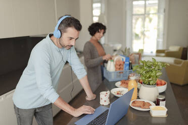 Mann mit Kopfhörern arbeitet von zu Hause aus am Laptop in der Küche - CAIF30174