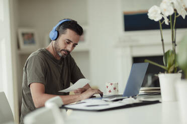 Man with headphones and receipts paying bills at laptop - CAIF30179