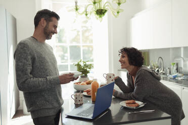 Glückliches Paar beim Frühstück und bei der Arbeit am Laptop in der Küche - CAIF30190