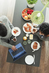 Couple enjoying breakfast and working at laptop in morning kitchen - CAIF30198