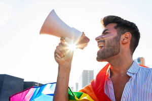 Seitenansicht eines exzentrischen schwulen Mannes mit Regenbogen-LGBT-Flagge, der in der Stadt steht und mit geschlossenen Augen in einen Lautsprecher schreit - ADSF20081