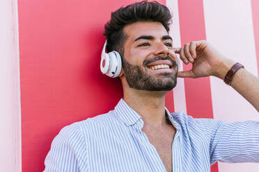 Carefree gay male with LGBT bracelet and in headphones standing on street on colorful wall while enjoying songs and looking away - ADSF20082