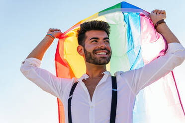 Low angle of expressive homosexual male standing on street with bright rainbow flag and cheerfully screaming - ADSF20083