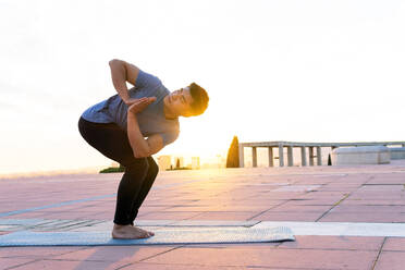 Seitenansicht eines flexiblen asiatischen Mannes in Parivrtta Utkatasana, der auf der Matte balanciert, während er bei Sonnenuntergang Yoga auf der Matte macht und nach oben schaut - ADSF20088