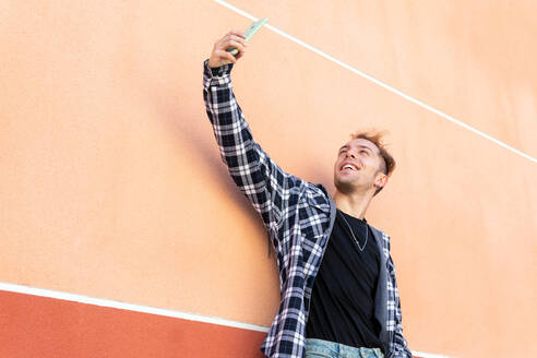 Niedriger Winkel des fröhlichen jungen Hipster-Mannes im karierten Hemd, der ein Selfie für soziale Medien auf dem Handy macht, während er an einer orangefarbenen Wand auf der Straße steht - ADSF20089