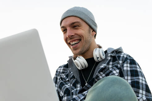 Niedriger Winkel von glücklichen jungen Hipster männlichen Blogger in lässigen Outfit mit Kopfhörern auf Hals sitzen auf Stein Grenze und Surfen Laptop bei Sonnenuntergang Zeit - ADSF20091