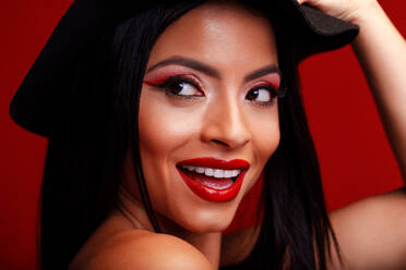 Fröhliche Frau mit roten Lippen und schwarzem Hut steht auf rotem Hintergrund und lächelt charmant, während sie den Kopf berührt und wegschaut - ADSF20096