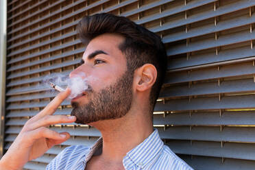 Side view of thoughtful handsome male leaning on building and smoking cigarette in city while looking away in contemplation - ADSF20135