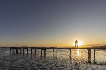 Silhouette einer Frau, die bei Sonnenaufgang allein auf einem Steg an der Küste steht - FOF11934