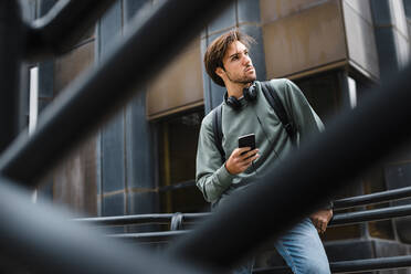 Male student with mobile phone looking away while leaning on railing at university campus - JMPF00781