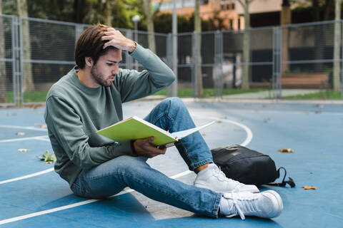 Junger männlicher Student, der ein Buch liest, während er auf einem Basketballplatz auf dem Universitätscampus sitzt, lizenzfreies Stockfoto