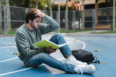 Junger männlicher Student, der ein Buch liest, während er auf einem Basketballplatz auf dem Universitätscampus sitzt - JMPF00782