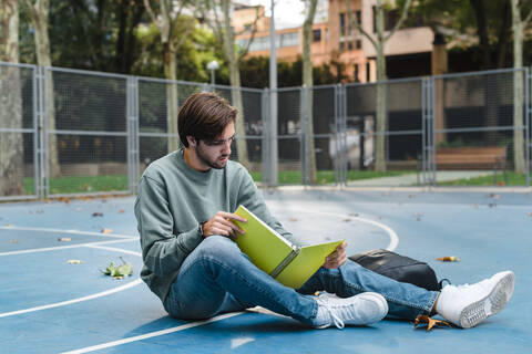 Junger Mann studiert beim Lesen eines Buches auf einem Basketballplatz in der Universität, lizenzfreies Stockfoto