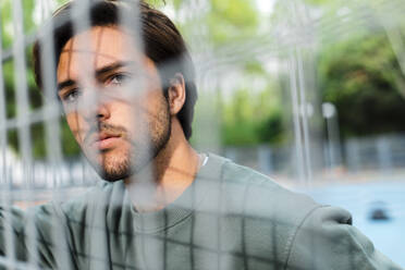 Thoughtful young handsome man looking away seen through fence at basketball court - JMPF00786