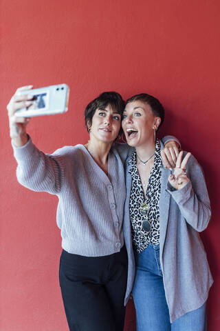 Junge Frau nimmt Selfie auf dem Handy mit aufgeregten Freund, während gegen rote Wand stehen, lizenzfreies Stockfoto