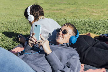 Lächelnde Frau mit Kopfhörern, die ein Mobiltelefon benutzt, während sie sich auf einen im Park liegenden Freund stützt - JRVF00147