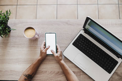 Geschäftsfrau benutzt Smartphone inmitten von Laptop und Kaffeetasse am Tisch - EBBF02209