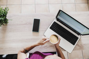 Businesswoman with laptop holding coffee cup on table at home office - EBBF02211