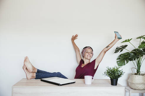 Relaxed businesswoman holding smart phone while looking away with feet up on table against wall - EBBF02216