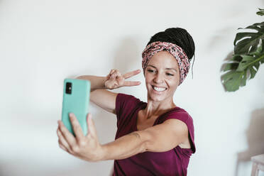 Lächelnde Frau, die ein Selfie mit ihrem Smartphone an der Wand zu Hause macht - EBBF02218