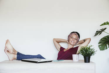 Smiling businesswoman relaxing with hands behind head against white wall at home office - EBBF02219