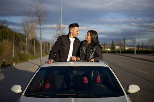 Junges Paar sieht sich an, während es auf dem Sonnendach eines Autos steht - OCMF01982