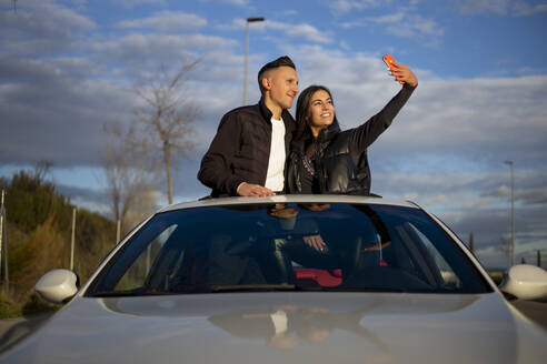 Lächelndes junges Paar, das ein Selfie mit dem Mobiltelefon macht, während es auf dem Sonnendach eines Autos steht - OCMF01984