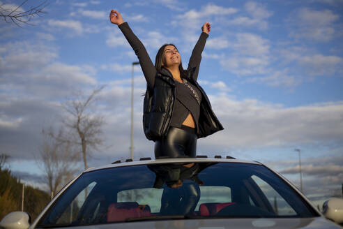 Glückliche Frau mit erhobener Hand im Auto stehend Sonnendach gegen Himmel - OCMF01986