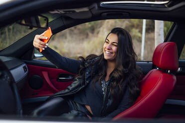 Lächelnde Frau, die ein Selfie mit ihrem Smartphone macht, während sie im Auto sitzt - OCMF01993