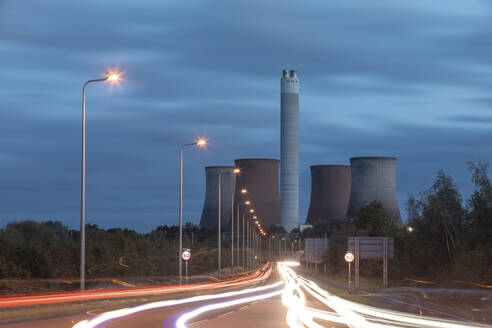 UK, England, Rugeley, Vehicle light trails stretching along illuminated road at dusk with cooling towers in background - WPEF03952