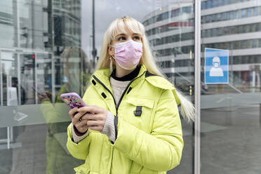 Junge Frau mit Gesichtsmaske, die ein Smartphone benutzt, während sie an einer Glaswand steht - FLLF00572