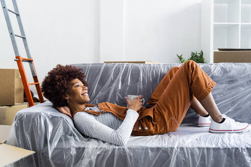 Smiling woman with coffee cup lying on sofa at new home - GIOF10710