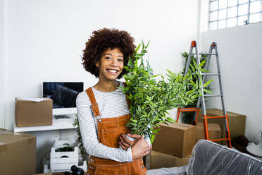 Happy afro woman holding potted plant during relocation in new apartment - GIOF10712