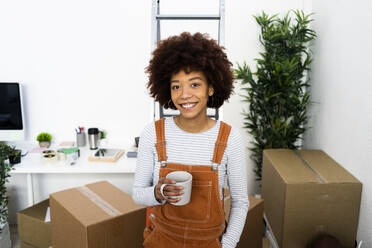 Glückliche junge Afro-Frau hält eine Kaffeetasse, während sie in einer neuen Loft-Wohnung steht - GIOF10715