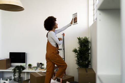 Afro-Frau, die einen Bilderrahmen an die Wand hängt, während sie auf eine Leiter in ihrer neuen Wohnung steigt - GIOF10716