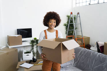 Smiling young holding cardboard box while standing in new home during relocation - GIOF10717