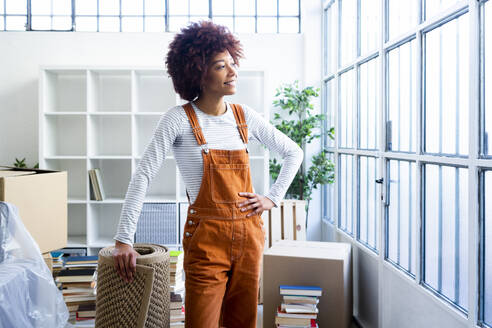 Lächelnde Afro-Frau mit Teppich, die durch ein Fenster in einer neuen Loft-Wohnung schaut - GIOF10720