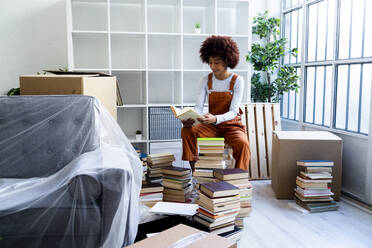 Afro woman reading book while unpacking during relocation in new home - GIOF10721