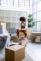 Afro-Frau packt Bücher aus, während sie an einem Regal in ihrer neuen Loft-Wohnung sitzt - GIOF10722