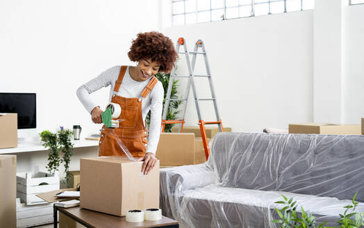 Afro woman packing box with adhesive tape while relocation in new home - GIOF10733