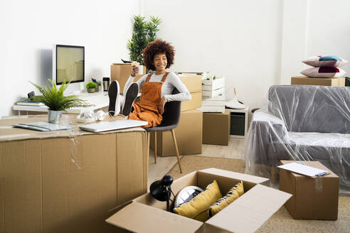 Afro woman looking away while holding coffee cup in living room at new home - GIOF10751