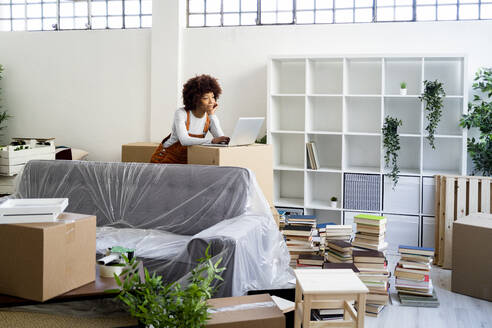 Afro-Frau mit Laptop, die sich beim Umzug in eine neue Wohnung auf einen Karton stützt - GIOF10754