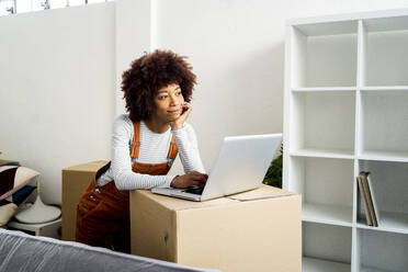 Afro woman with hand on chin day dreaming while standing by laptop in new loft apartment - GIOF10755