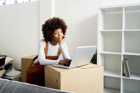 Afro-Frau mit Hand am Kinn träumt, während sie am Laptop in einer neuen Loft-Wohnung steht - GIOF10755