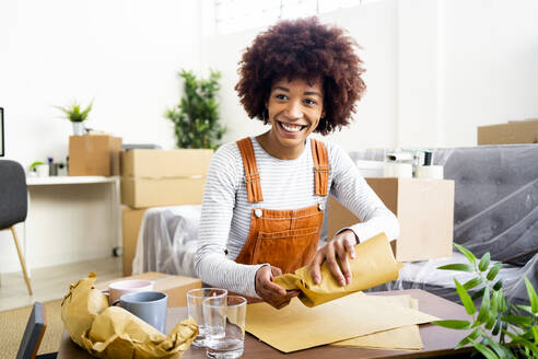 Happy Afro young woman wrapping drinking glass in brown paper at new home - GIOF10760