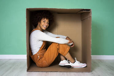 Lächelnde Afro-Frau, die in einem Karton in ihrem neuen Zuhause sitzt - GIOF10765