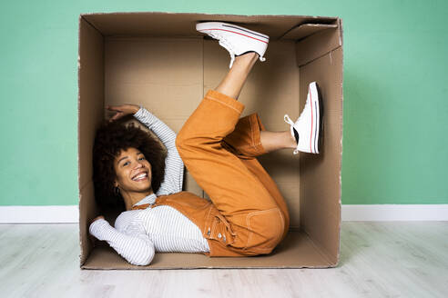 Cheerful woman lying in cardboard box against green wall in new loft apartment - GIOF10766