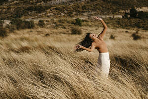 Junge Frau mit erhobenen Armen steht inmitten von Gras auf einem Feld an einem windigen Tag - TCEF01434