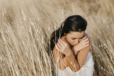 Nachdenkliche junge Frau sitzt bei Sonnenuntergang im Gras auf einem Feld - TCEF01437