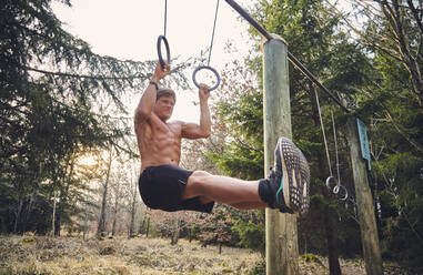 Männlicher Athlet ohne Hemd, der auf einem Fitnessparcours im Wald an Gymnastikringen hängt - KDF00740